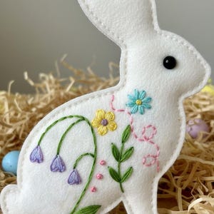 May include: White felt bunny decoration with embroidered flowers in yellow, blue, and purple. The bunny has a black eye and a pastel rainbow ribbon for hanging. Easter decor.