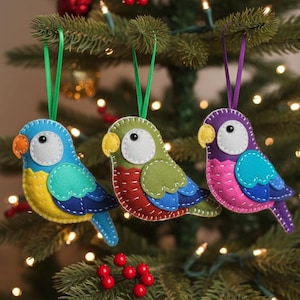 May include: Three colorful felt parrot ornaments hanging on a Christmas tree. The parrots are blue, green, and purple, with white, yellow, and orange accents. Each ornament has a green or purple ribbon for hanging.