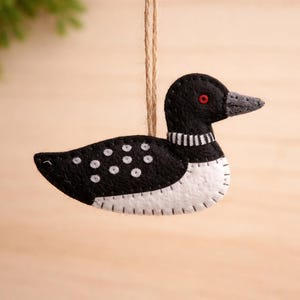 May include: A handmade felt loon decoration. The bird is black and white with a grey beak and red eye. It hangs from a natural twine string. The loon has white spots on its back and a white belly.
