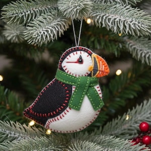 Peut inclure: Ornement de Noël en feutre représentant un macareux. Le dos est noir, le ventre blanc, et le bec orange. Il porte une écharpe verte et des coutures rouges. Suspendu à un fil argenté sur un sapin de Noël.