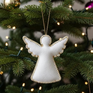 Könnte beinhalten: Ein weißer Weihnachtsbaumschmuck in Form eines Engels aus Filz mit goldfarbenen Nähten und einem goldenen Aufhängeband. Der Engel hat ausgebreitete Flügel und hängt an einem Weihnachtsbaum mit Lichtern.