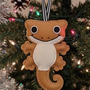May include: A brown felt gecko Christmas ornament with a white belly, black eyes, and a smiling face. The ornament is hanging from a black and white striped ribbon against a backdrop of a decorated Christmas tree.