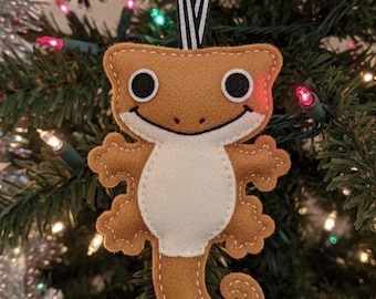 May include: A brown felt gecko Christmas ornament with a white belly, black eyes, and a smiling face. The ornament is hanging from a black and white striped ribbon against a backdrop of a decorated Christmas tree.