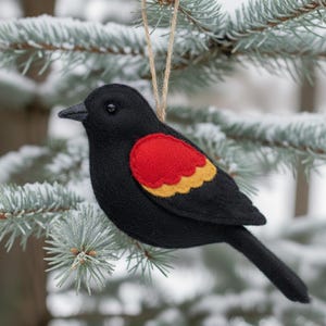 May include: A felt bird ornament with a black body, a red and yellow wing patch, and a jute string for hanging. The bird is perched on a snowy evergreen branch, creating a festive holiday decoration.