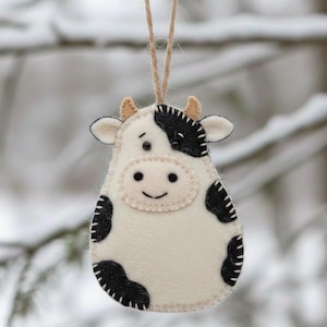 Puede incluir: Adorno de vaca de fieltro crema con manchas negras, una cara sonriente y pequeños cuernos marrones. El adorno tiene un contorno cosido y cuelga de una cuerda de hilo natural. Ideal para decoraciones navideñas.
