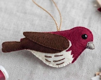 Puede incluir: Un adorno de pájaro de fieltro con cabeza y espalda de color rojo oscuro, alas marrones y vientre blanco con detalles cosidos. El pájaro tiene un pequeño ojo de cuenta negra, un pico gris claro y un lazo de cuerda marrón para colgarlo.
