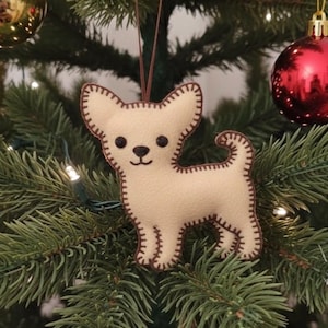 May include: A beige felt Chihuahua dog Christmas tree ornament with brown stitched details. The ornament has a brown hanging loop and is displayed on a Christmas tree with other ornaments and lights.