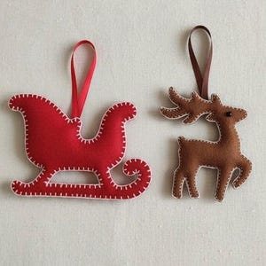 May include: Two felt Christmas ornaments: a red sleigh with a white stitched outline and a brown reindeer with a white stitched outline. Both have ribbon hangers.