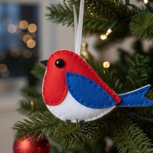 May include: A felt bird Christmas ornament with a red head and chest, blue wing, and white belly. The bird is hanging on a green Christmas tree with a white ribbon. A red ornament is also visible.