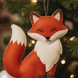 Puede incluir: Adorno navideño de fieltro con forma de zorro. El adorno es principalmente naranja con pecho y punta de la cola blancos. Tiene detalles marrones en las orejas y patas. El zorro sonríe y cuelga de un hilo dorado, sobre un árbol de Navidad.