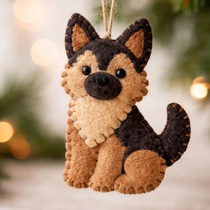 Könnte beinhalten: Ein Filz-Ornament in Form eines Deutschen Schäferhundes. Der Hund hat einen braunen Körper mit schwarzen und braunen Markierungen, schwarze Augen und eine schwarze Nase. Eine Schnurschlaufe ist zum Aufhängen befestigt. Weihnachtsdekoration.