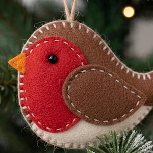 May include: A felt Christmas ornament shaped like a robin bird. The bird has a red breast, brown wings, and a beige body. The ornament is decorated with white stitching and an orange beak. A string is attached for hanging.