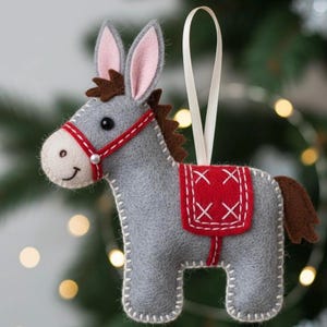 May include: A gray felt donkey Christmas ornament with a red saddle and bridle. The donkey has brown hair, pink ears, and a white ribbon for hanging. The ornament is stitched with white thread.