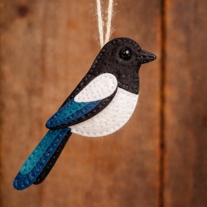 May include: A handmade felt bird ornament in black, white, and teal blue, suspended by a natural twine string. The bird features a black head and beak, white body, and teal blue tail and wing accents. A decorative item.