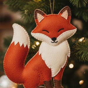 May include: A felt fox Christmas ornament with a white chest and tail, brown ears and feet, and a smiling face. The fox is hanging from a gold string against a backdrop of a Christmas tree with lights and ornaments.