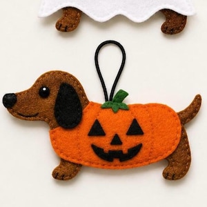 May include: A brown felt dachshund ornament wearing an orange pumpkin costume with a black jack-o'-lantern face. The dog has a black nose, black ears, and a black elastic loop for hanging. A white felt ghost shape is visible at the top edge.
