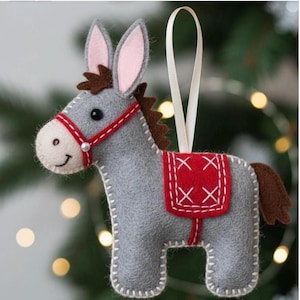 May include: A grey felt donkey ornament featuring a cream-coloured muzzle, pink-lined ears, and a brown mane and tail. The donkey wears a red felt bridle with a small white bead and a red saddle blanket with white cross-stitch detailing and a white ribbon.