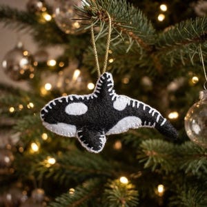 May include: A handcrafted felt orca ornament featuring a black and white body with visible white whip-stitching along the edges. The whale-shaped decoration is suspended by a thin gold cord, showcasing a dorsal fin and distinct eye patch.