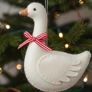 May include: A white felt goose ornament featuring a tan beak and a red and white striped ribbon tied in a bow around its neck. The goose has visible stitching along the edges of its body and wing, and a thin string attached for hanging purposes.