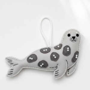 Könnte beinhalten: Graues Filz-Seehund-Ornament mit dunkelgrauen Flecken und weißen Nähten. Der Seehund hat schwarze Augen, eine schwarze Nase und kleine weiße Perlenakzente. Zum Aufhängen ist eine weiße Schnur befestigt.