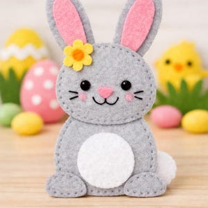 May include: A grey felt bunny with pink inner ears and a yellow flower. The bunny has a white belly and tail, black eyes, and a pink nose. Easter decorations are in the background.