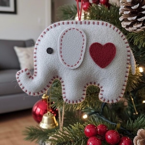 May include: A felt elephant Christmas ornament with a red heart and stitched details. The gray elephant has a black button eye and is hanging on a decorated Christmas tree with red berries and pinecones.