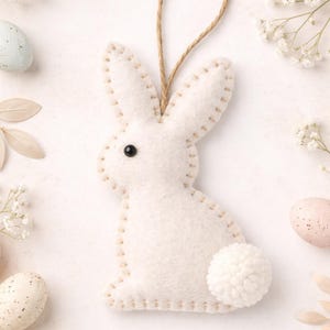 Puede incluir: Adorno de conejito de fieltro blanco con un contorno cosido y una cola de pompón. El conejito tiene una cuerda de yute para colgar y un ojo negro. La imagen está rodeada de huevos de Pascua pastel y flores blancas.