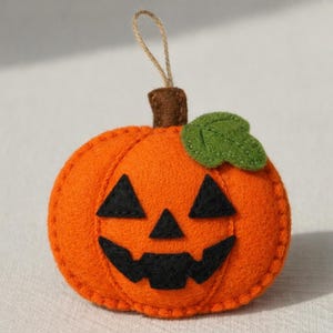 Può includere: Ornamento a forma di zucca in feltro con un volto sorridente di Jack-o'-lantern. La zucca arancione ha occhi e bocca triangolari neri, una foglia verde e un gambo marrone. Un anello di spago è attaccato per appenderlo. Decorazione di Halloween.