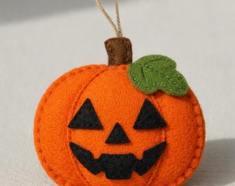 May include: A felt pumpkin ornament with a smiling jack-o'-lantern face. The orange pumpkin has black triangle eyes and a mouth, a green leaf, and a brown stem. A loop of twine is attached for hanging. A festive Halloween decoration.