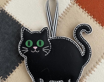 May include: A black cat ornament with green eyes and a pink nose, outlined with white stitching. It has a striped black and white ribbon for hanging. The ornament is on a patchwork background.