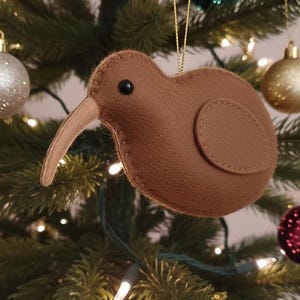 Könnte beinhalten: Ein brauner Filz-Kiwi-Vogel-Weihnachtsschmuck mit langem Schnabel und kleinem, rundem Flügeldetaill. Der Schmuck hängt an einer goldenen Schnur vor dem Hintergrund eines geschmückten Weihnachtsbaums mit bunten Kugeln und Lichtern.