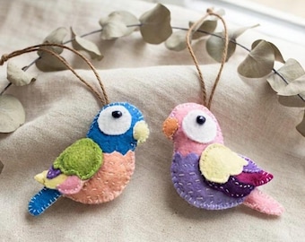 Puede incluir: Dos adornos de pájaros de fieltro hechos a mano con lazos de cuerda para colgar. El pájaro de la izquierda presenta fieltro azul, verde, melocotón y amarillo con ojos de cuentas negras. El de la derecha tiene fieltro rosa, morado, amarillo y naranja.