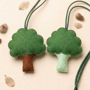 May include: Two felt tree ornaments. One has a brown trunk and green leaves, the other has a light green trunk and green leaves. Both are suspended by green cords. The background is a light beige color with small pebbles and a leaf.