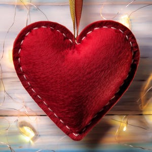 Puede incluir: Un adorno en forma de corazón de felpa, en un tono rojo vibrante. El corazón está cosido con hilo blanco alrededor de los bordes y tiene una superficie texturizada. Se adjunta una cinta dorada y roja para colgarlo. Luces decorativas en el fondo.
