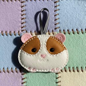 May include: A handmade felt hamster ornament with brown, white, and pink details. The hamster has black eyes, a pink nose, and pink ears. It is stitched with white thread and has a black and white striped ribbon for hanging.