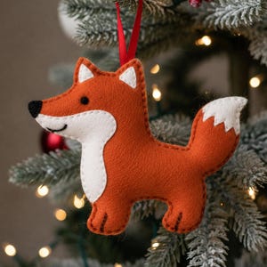 May include: A festive felt fox Christmas decoration. The fox is a vibrant orange with white detailing on its chest, ears, and tail. It features a black nose and a red ribbon for hanging. The ornament hangs on a Christmas tree.