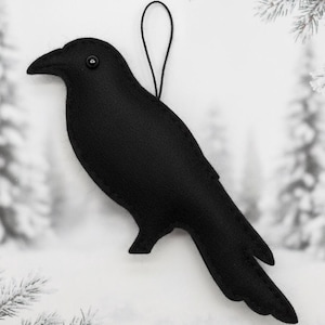 Puede incluir: Adorno de cuervo negro de fieltro con un botón como ojo y una cuerda negra para colgar. El pájaro tiene un pico y plumas de la cola detallados. El fondo es una escena invernal borrosa.