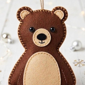 May include: A handmade brown felt bear ornament with a tan belly and ears. The bear has black eyes and a tan nose. The edges are stitched with tan thread, and a twine loop is attached for hanging. Decorative holiday item.