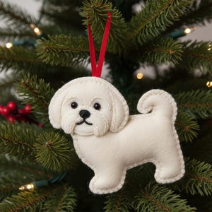 Puede incluir: Adorno navideño de fieltro blanco con forma de perro y cinta roja para colgar. El perro tiene ojos negros y nariz negra. El adorno cuelga de un árbol de Navidad verde.