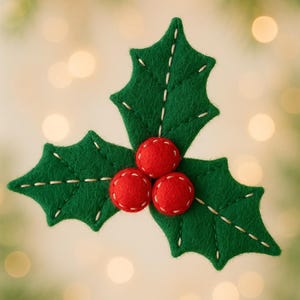 Op de afbeelding: Een handgemaakte kerstversiering met hulstmotief. De afbeelding toont drie groene viltbladeren met witte stiksels en drie rode viltbessen. De achtergrond is wazig met zachte, onscherpe lichten.