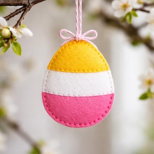 May include: A felt Easter egg ornament in yellow, white, and pink, hanging from a pink string tied in a bow. The egg is suspended from a flowering tree branch, creating a spring-themed decoration.