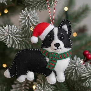 Puede incluir: Adorno navideño de fieltro con forma de perro blanco y negro, con gorro de Papá Noel rojo y bufanda verde con detalles rojos. El adorno cuelga de una cuerda roja y blanca en un árbol de Navidad.