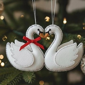 Puede incluir: Dos adornos navideños de cisnes blancos, con detalles cosidos en negro y un lazo rojo. Los cisnes están colocados para tocarse los picos. El fondo es un árbol de Navidad borroso con luces.
