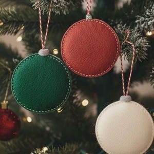 May include: Close-up of a Christmas tree adorned with three round ornaments. The ornaments are in green, red, and white, with white stitching and striped hanging strings. The tree has green needles and small lights.