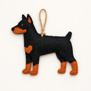 May include: A felt dog ornament in a standing pose, featuring a black body with orange accents on the ears, snout, and legs. A small orange heart is on the side. A twine loop is attached for hanging.