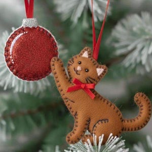 Può includere: Due decorazioni natalizie appese a un albero. Una è una palla rossa glitterata con una parte superiore argentata. L'altra è un gatto di feltro marrone con un fiocco rosso, dettagli bianchi e cuciture. Entrambi hanno nastri rossi.