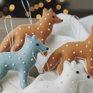 Può includere: Primo piano di quattro ornamenti a forma di volpe in feltro in vari colori: azzurro, bianco e due tonalità di marrone. Ogni volpe è decorata con piccole perline bianche a forma di perla e ha un filo d'argento per appenderla. Lo sfondo è sfocato con luci natalizie.