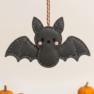 Könnte beinhalten: Handgefertigter Fledermaus-Anhänger aus dunkelgrauem Filz mit weißen Nähten. Die Fledermaus hat rosa Wangen und eine schwarz-orangefarbene Schnur zum Aufhängen. Geeignet als Dekoration für Halloween oder die Herbstsaison.