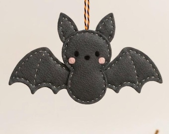 May include: A handmade felt bat ornament in dark gray with white stitching. The bat has pink cheeks and a black string for hanging. The wings are spread, and the ornament is designed for Halloween or seasonal decor.