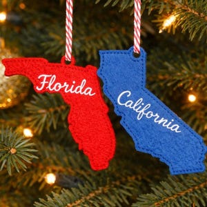 May include: Red and blue felt Christmas ornaments shaped like the states of Florida and California. Each ornament has the state name embroidered in white. They hang from red and white striped cords, displayed on a Christmas tree.